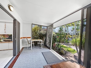 A balcony with a table and chairs, a TV, and a view of a garden and buildings. - Canopy Chalet 6 (Nelly Bay)