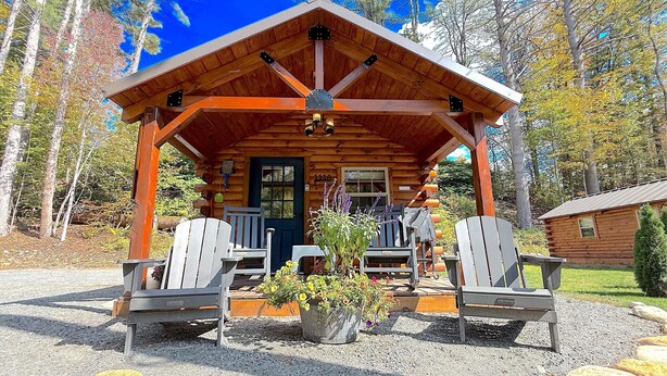 A log cabin with a porch, two Adirondack chairs, a potted plant, and a gravel driveway.