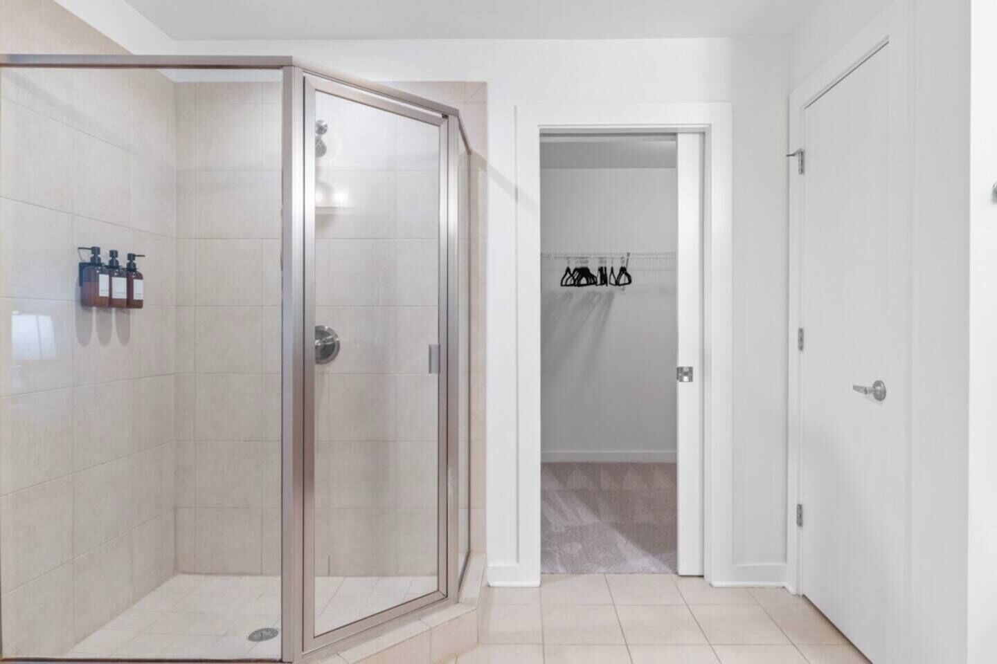 A modern bathroom with a glass-enclosed shower, white tiled walls, and a door leading to another room.