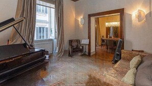 Een ruime kamer met een vleugelpiano, een bank en een eethoek met een kroonluchter.