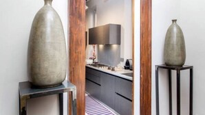 Een hal met twee grote vazen op voetstukken, een houten deurkozijn en een blik op een moderne keuken met donkere kasten en een aanrecht.