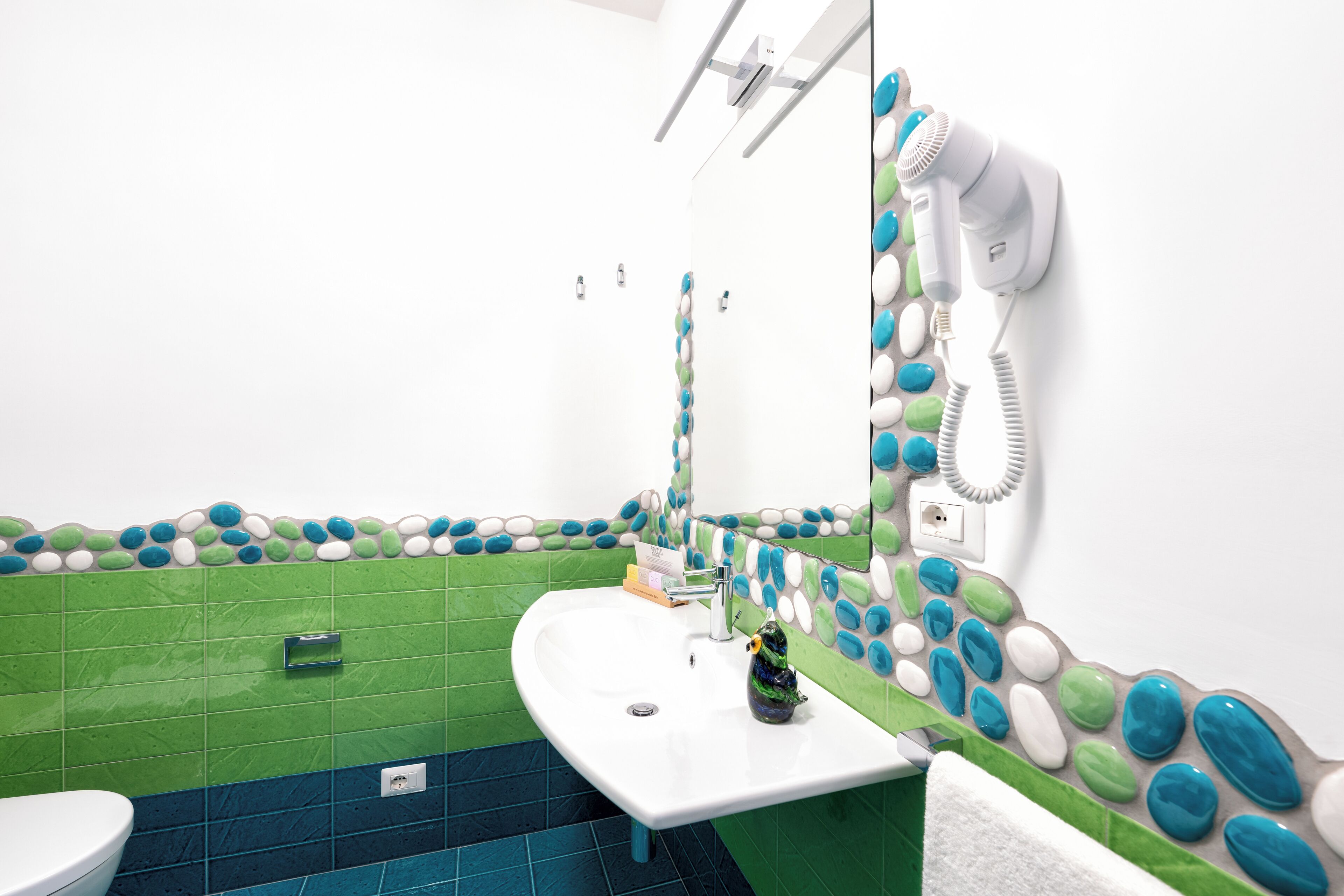 A bathroom with a green and white tiled wall, a white sink, and a hairdryer mounted on the wall.