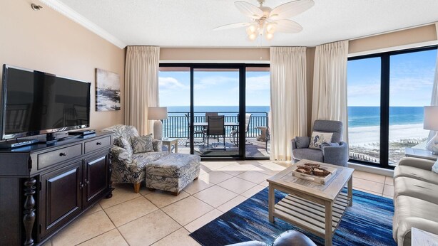 A spacious living room with a large TV, comfortable seating, a balcony with a view of the ocean, and a ceiling fan. - Seachase 704w (Orange Beach)