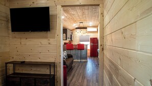 Een houten blokhut interieur met een aan de muur bevestigde televisie, een bank, een kleine tafel en een keuken met een koelkast en rode stoelen.