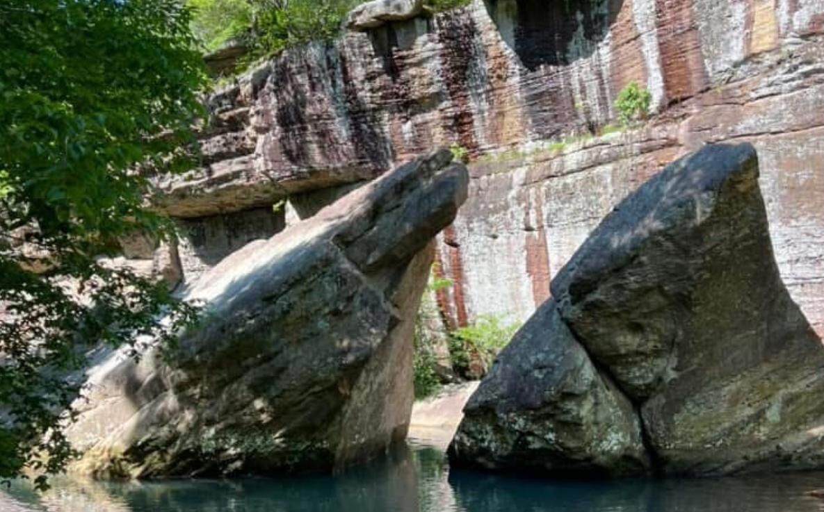 Formación rocosa natural con grandes rocas parcialmente sumergidas en agua, rodeada de vegetación.