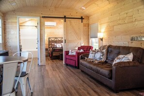Interior acogedor de una cabaña con piso de madera, un sofá de cuero marrón, una silla roja, una mesa de comedor con sillas y una cama con estructura de metal.