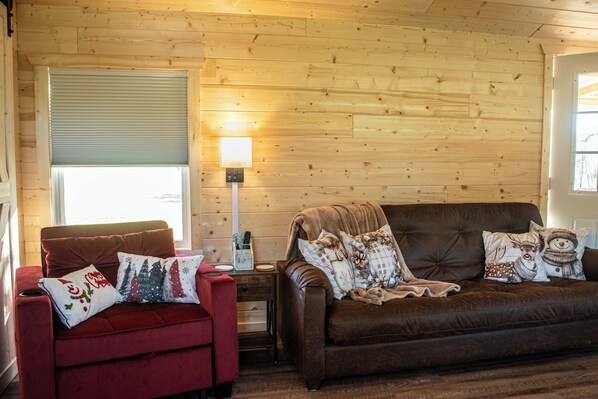 A cozy living room with a red armchair and a brown sofa, both adorned with festive pillows. A wooden wall and a window with blinds are visible.