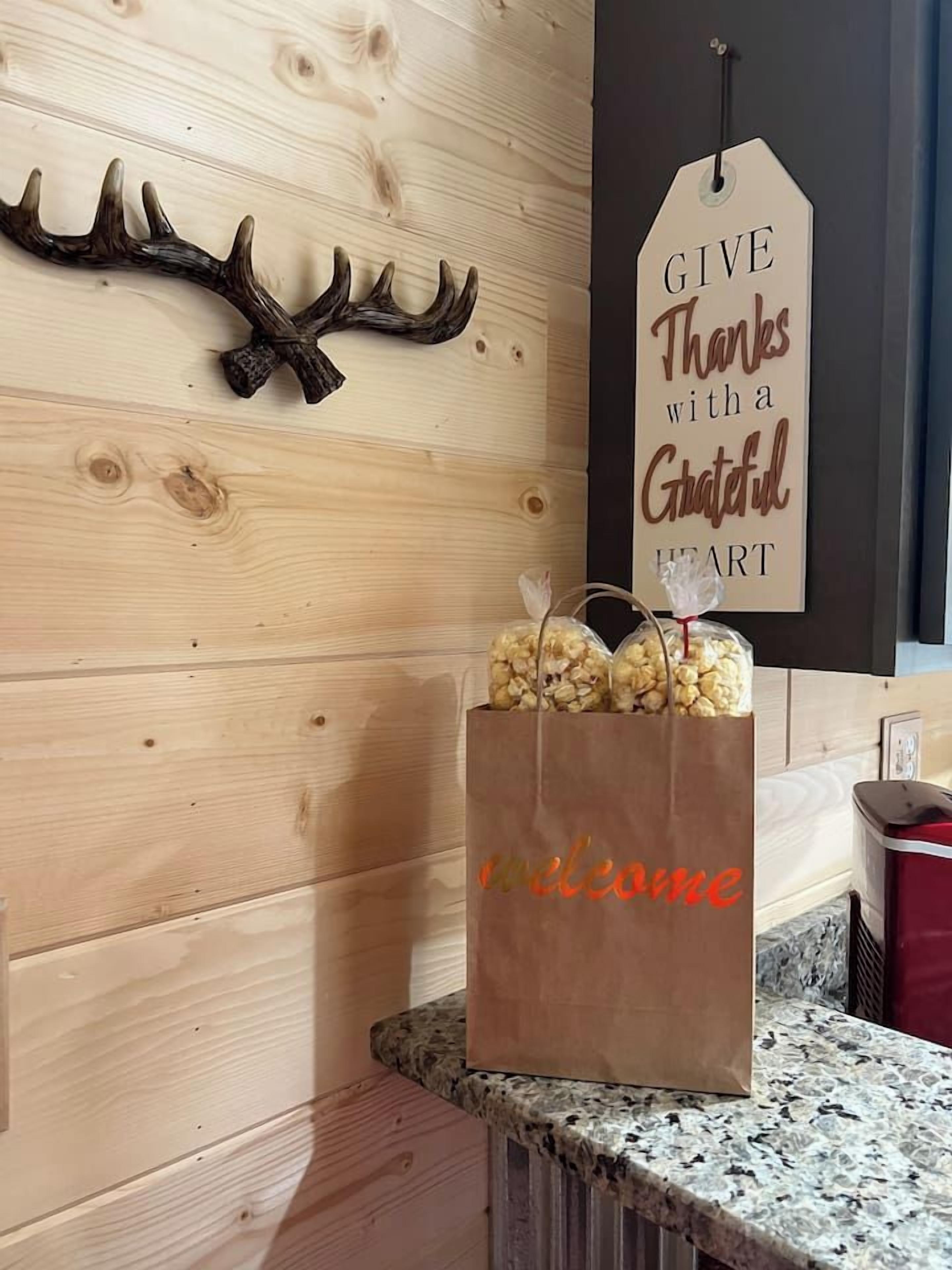 Pared de madera con una decoración de cornamenta de ciervo y un letrero que dice 'GIVE Thanks with a Grateful Heart'. Una bolsa de papel marrón con la palabra 'welcome' escrita está sobre una encimera de granito.