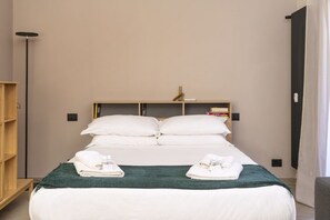 A hotel room with a bed, pillows, a bedside table with books, a floor lamp, and a wardrobe.
