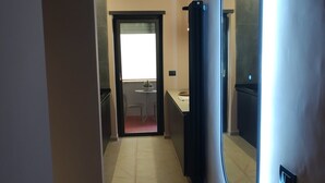 Um corredor com piso ladrilhado, uma porta que dá para um quarto e um espelho com iluminação na parede.