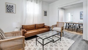 A living room with a brown leather sofa, a glass coffee table, wicker chairs, and a dining area with a wooden table and chairs. - Charming 2-bedroom house with AC, WiFi in wonderful Richmond (Richmond)
