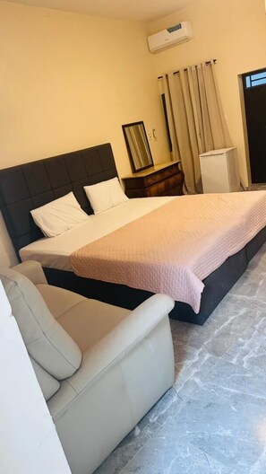 Ett hotellrum med en säng, en stol, en byrå, en luftkonditioneringsanläggning och ett fönster med gardiner.