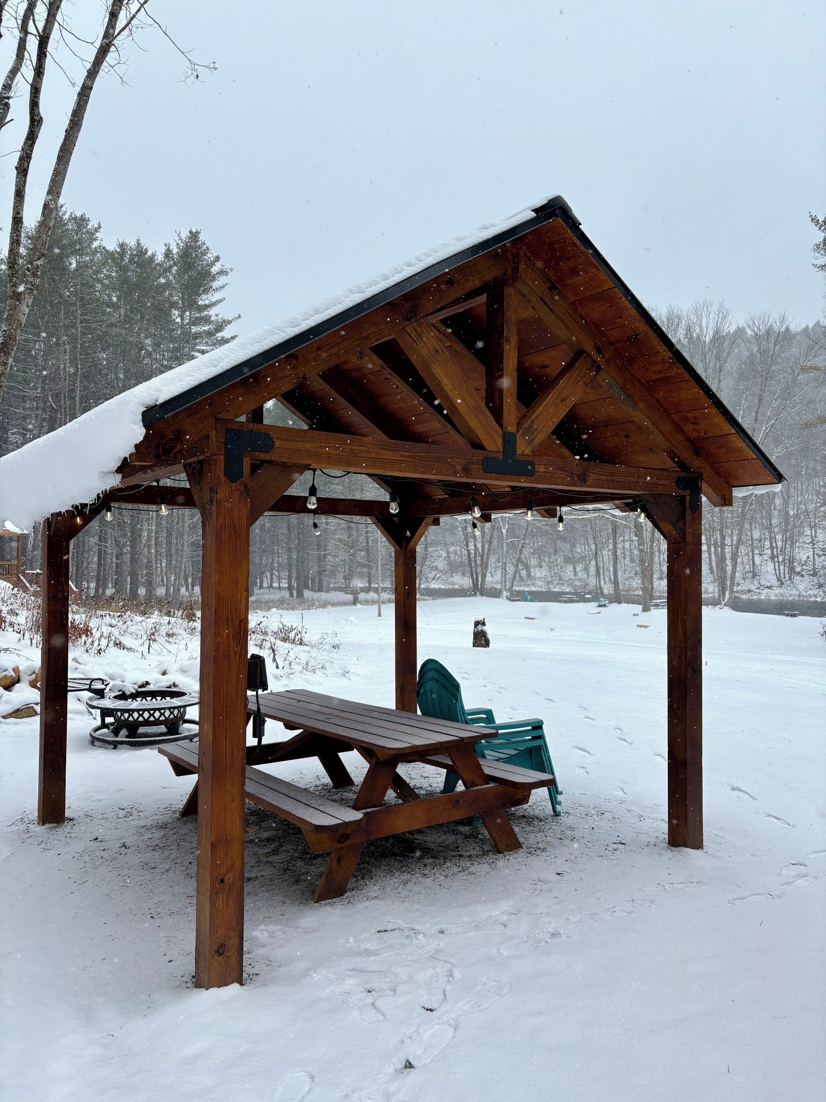 Un gazebo in legno con panchina e tavolo, immerso in un paesaggio innevato tra alberi.