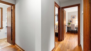 A hallway with wooden flooring and white walls, leading to a room with a desk and chair.