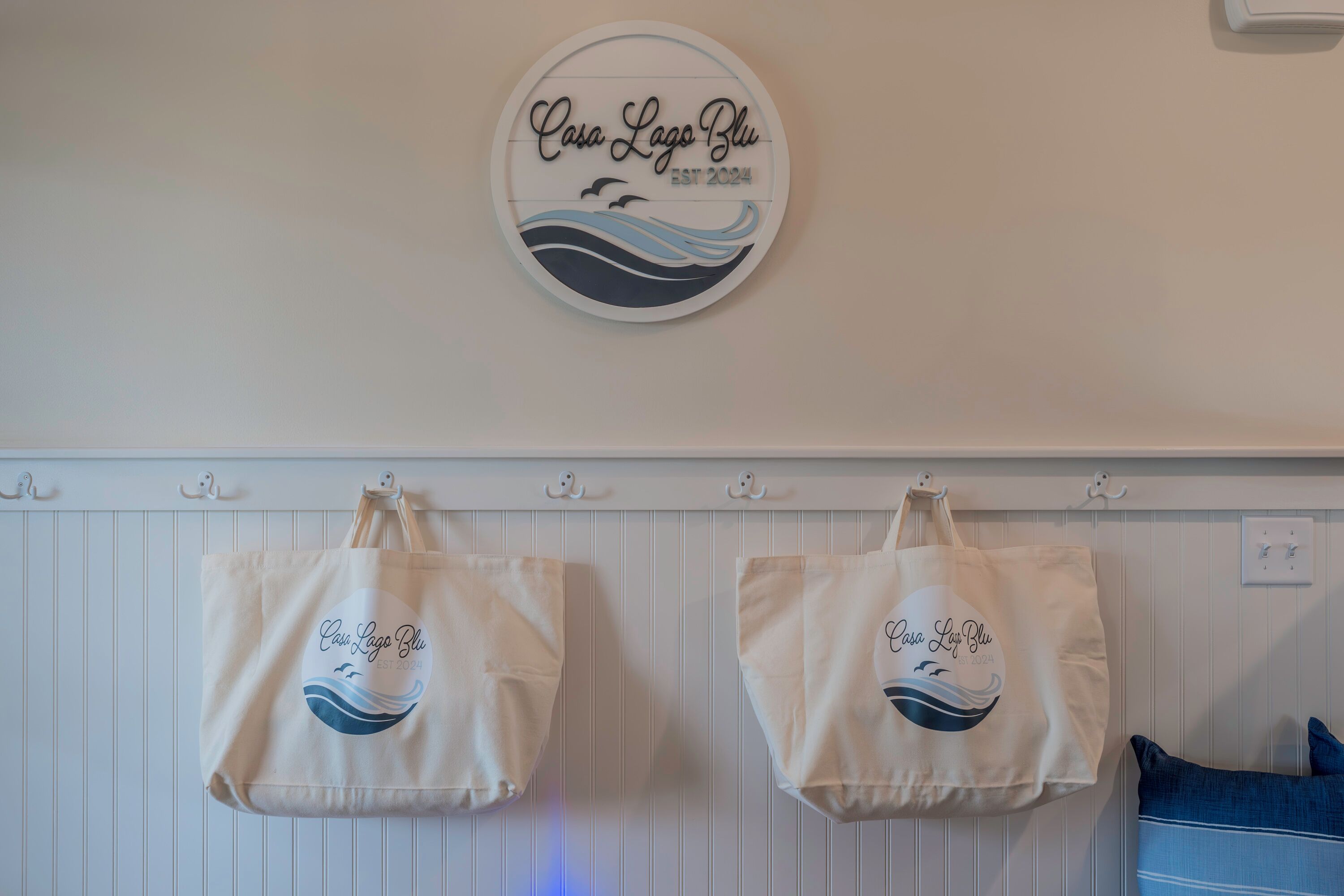 A wall with a circular sign reading "Casa Lago Blu" and two hanging tote bags with matching designs.