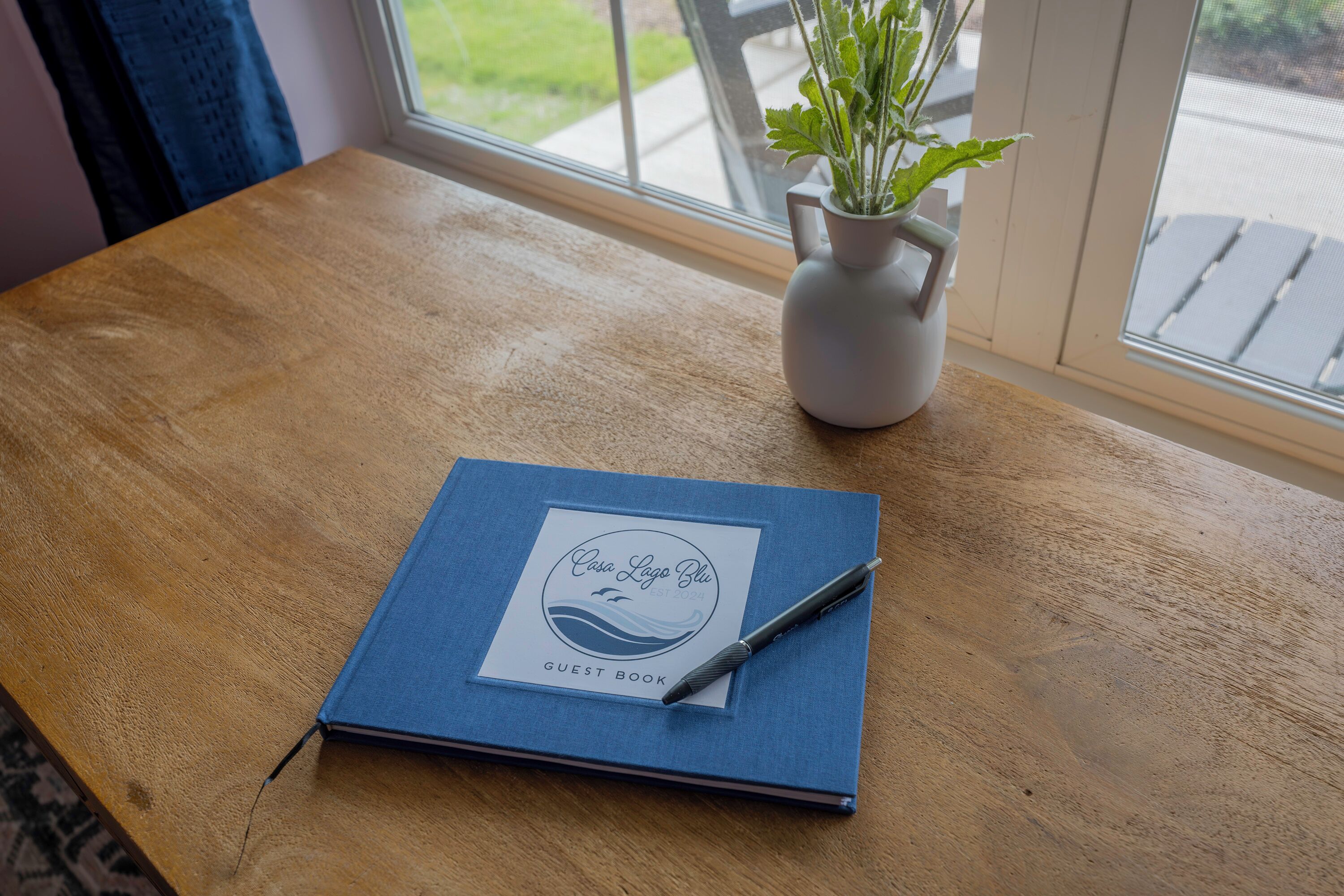 A blue guest book on a wooden table with a pen, next to a white vase with green plants.