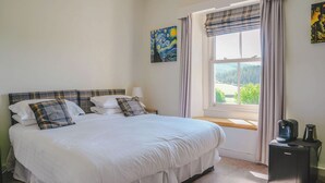 Een netjes opgemaakt bed met witte beddengoed en geruite kussens, een nachtkastje met een lamp, een raam met gordijnen en een klein zwart apparaat op de vloer.