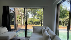 A modern living room with a large sofa, a coffee table, and a view of a swimming pool and garden through large windows. - Bryanston Luxurious 4-Bed Villa (Sandton)