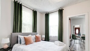 A modern bedroom with a large bed, green curtains, a bedside table with a lamp, and a view into a dining area.