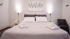 A neatly made bed with white linens, two bedside lamps, a wall-mounted air conditioner, and a framed artwork above the bed.