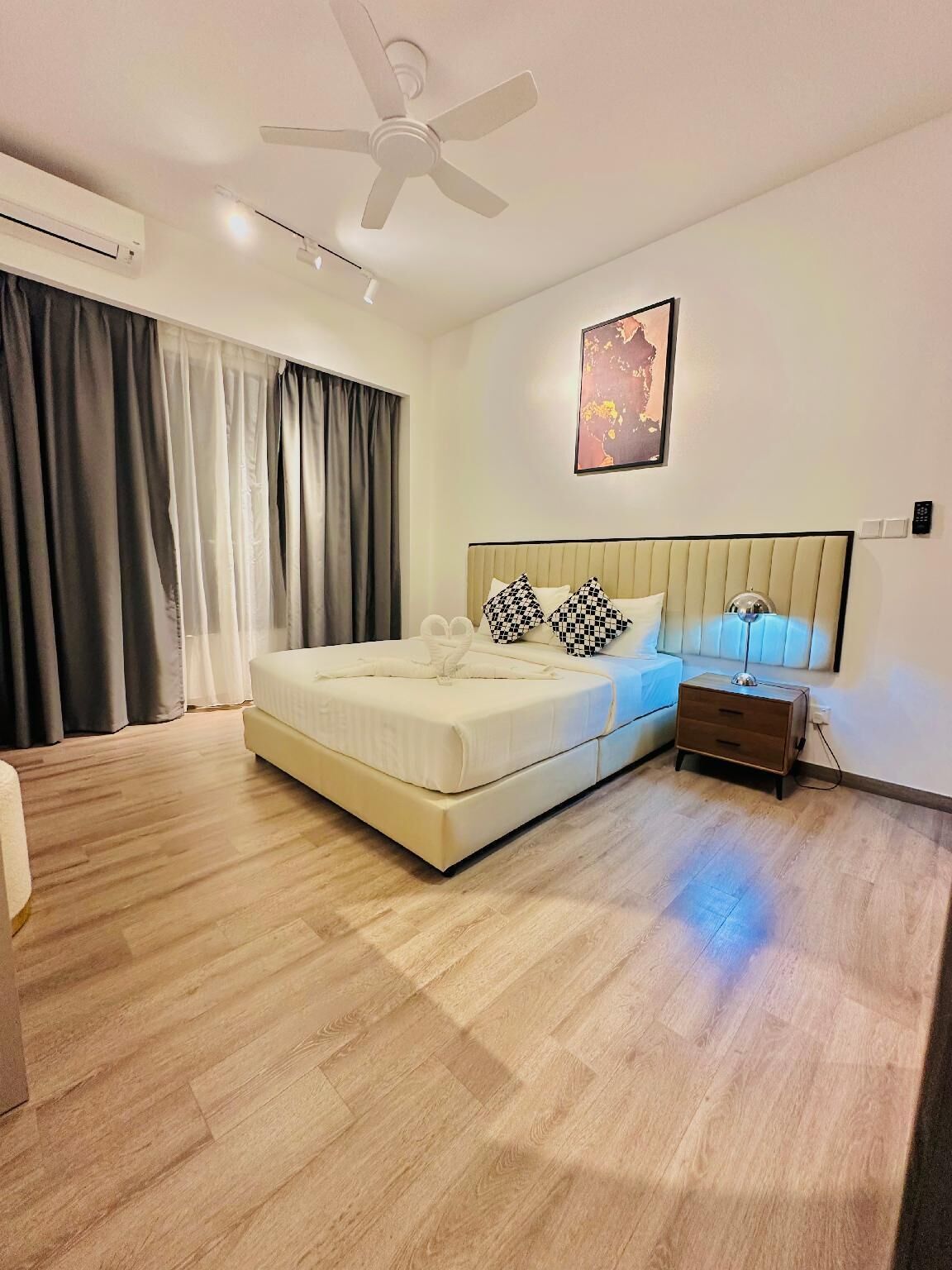 A modern bedroom with a large bed, a ceiling fan, an air conditioning unit, a bedside table with a lamp, and a painting on the wall.