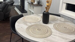 Uma área de jantar com uma mesa redonda, cadeiras pretas e jogos americanos trançados.