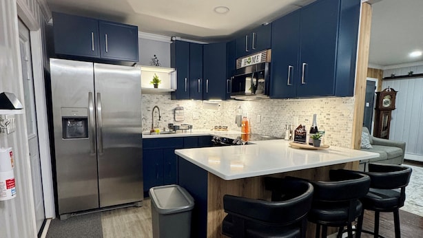 Uma cozinha moderna com armários azul escuro, uma geladeira de aço inoxidável, uma ilha central com banquetas e piso de madeira.