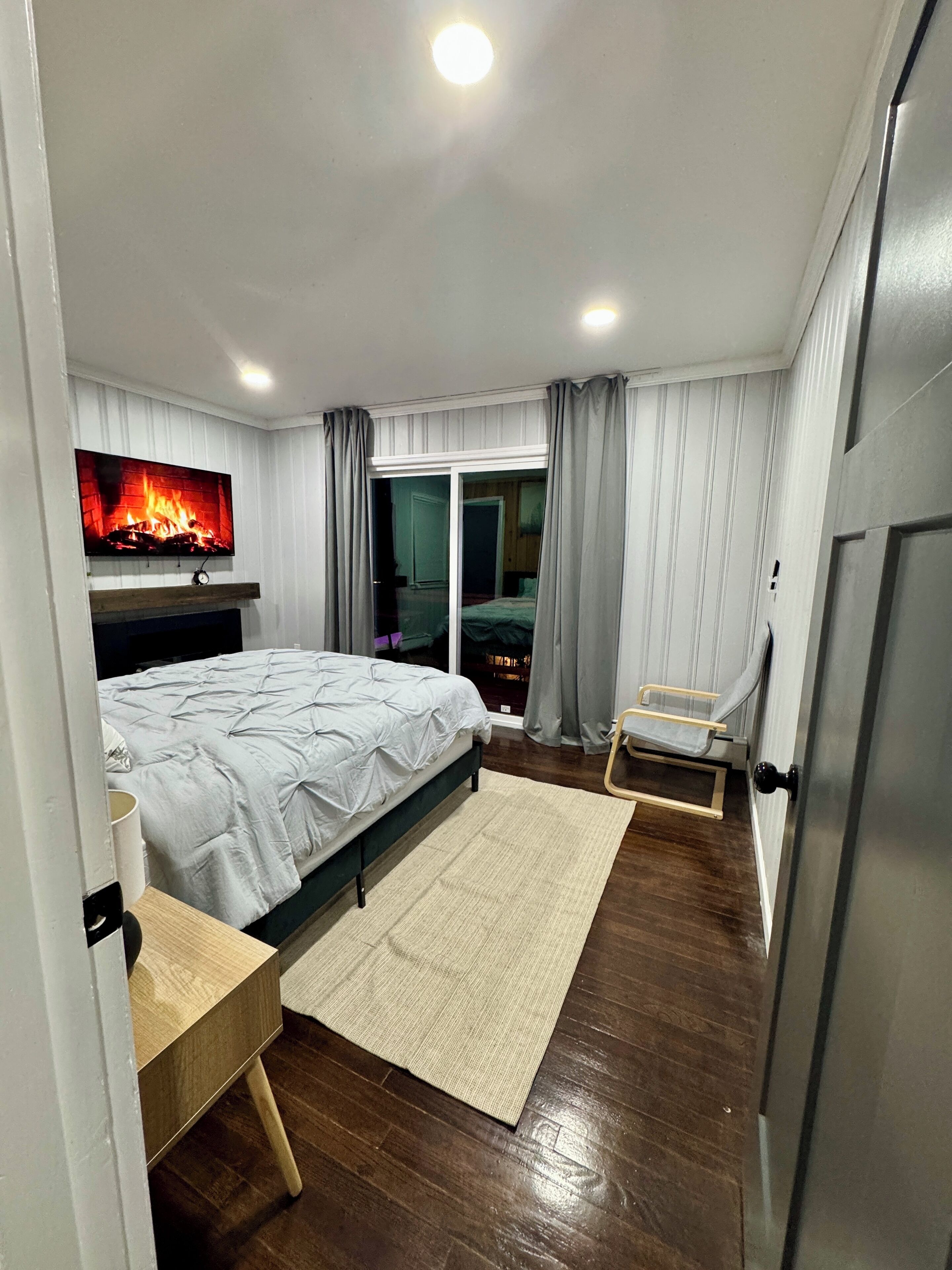 Une chambre moderne avec un lit, un téléviseur fixé au mur, une petite table et une porte coulissante donnant sur un balcon.