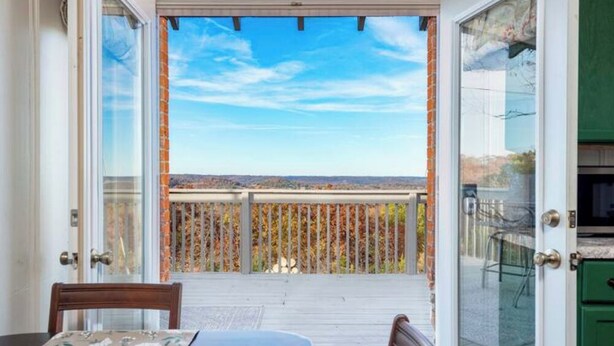 Un balcon offrant une vue sur un paysage lointain, une terrasse en bois et une porte vitrée donnant sur une pièce avec des meubles de couleur verte.