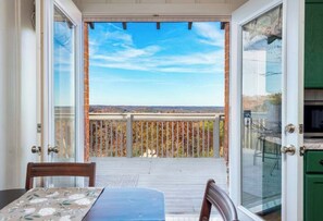 Un balcon offrant une vue sur un paysage lointain, une terrasse en bois et une porte vitrée donnant sur une pièce avec des meubles de couleur verte.