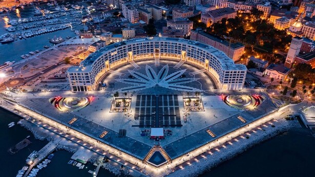 Vue aérienne d’une promenade en bord de mer, avec une place circulaire, des bateaux amarrés le long du rivage et des bâtiments environnants.