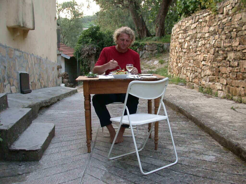 A person sitting at a wooden table outdoors with a white chair, a stone wall, and a stone staircase in the background.