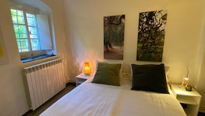 A bedroom with a large bed, a window, a radiator, and two wall-mounted paintings. - Ligurian rustico, surrounded by olive groves, 10 minutes from the sea. (Imperia)