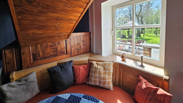 Una habitación acogedora con techo de madera, un banco con cojines y una ventana con vistas a un jardín.