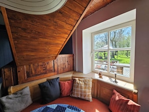 Ein gemütliches Zimmer mit Holzbalkendecke, einer Bank mit Kissen und einem Fenster mit Blick auf einen Garten.