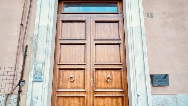 Puerta doble de madera con un tirador metálico, un ventanal arriba y el número '25' en la pared a la derecha.