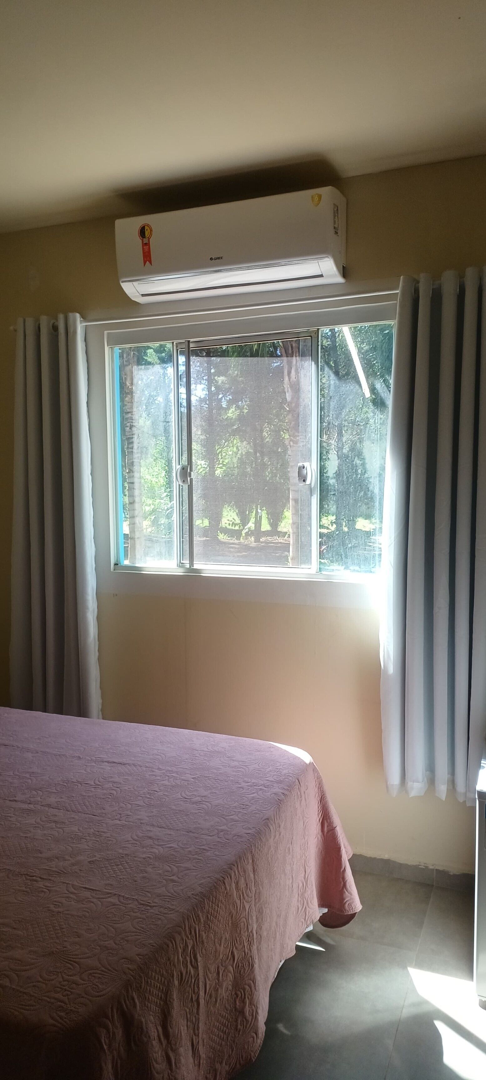 A room with a bed, an air conditioning unit, a window with curtains, and a view of greenery outside.