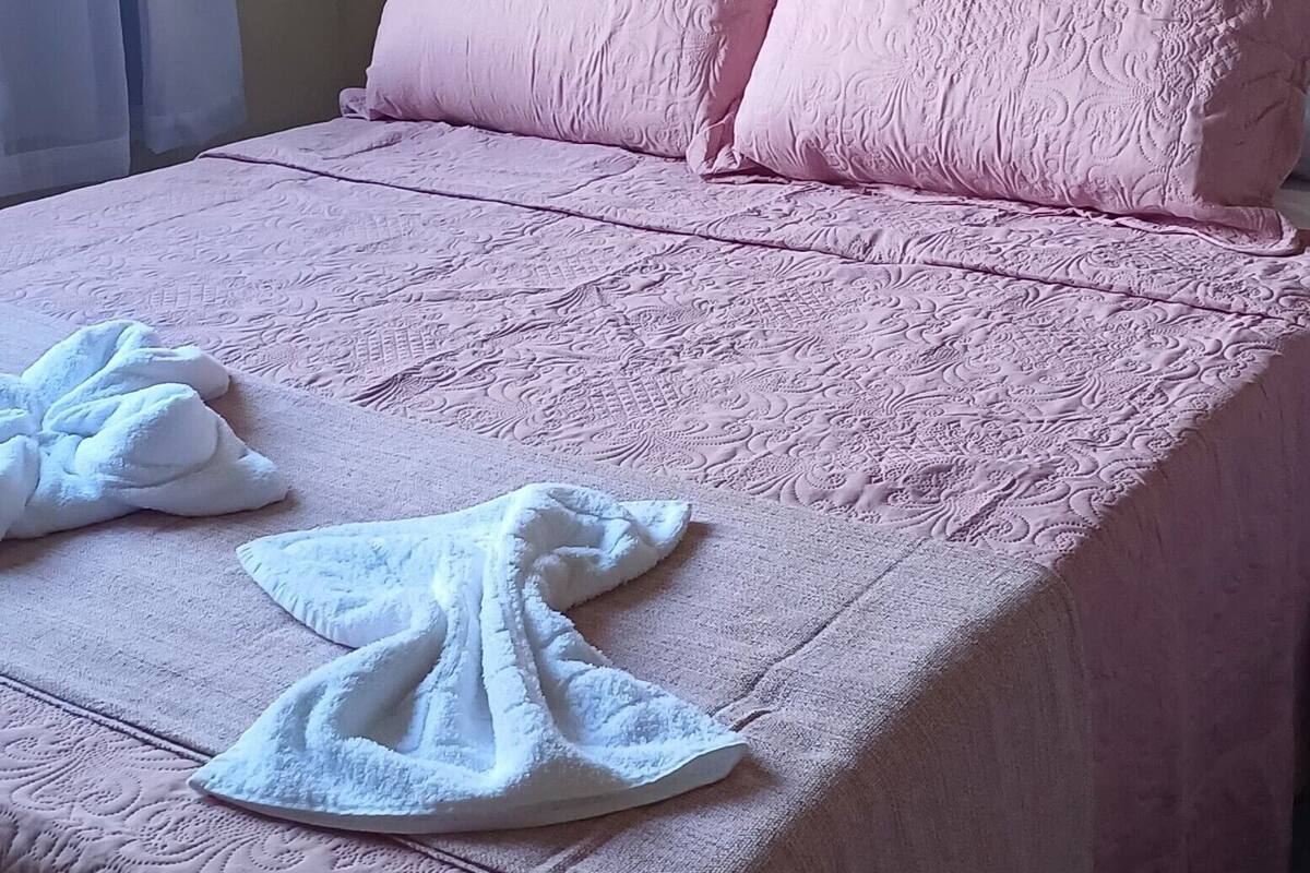 Un lit avec une couvre-lit rose et deux oreillers, une serviette blanche et une couverture blanche sur le lit.