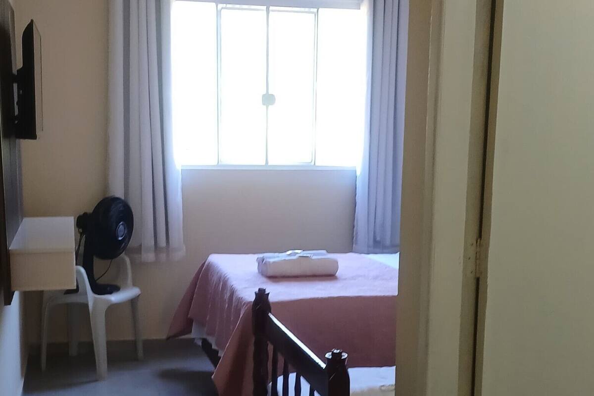 Une pièce comprenant un lit, un bureau, une chaise, un ventilateur et une fenêtre avec des rideaux.