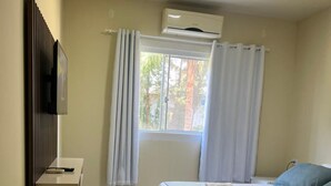 Une chambre à coucher comprenant un lit, un téléviseur fixé au mur, une fenêtre avec des rideaux et un appareil de climatisation.