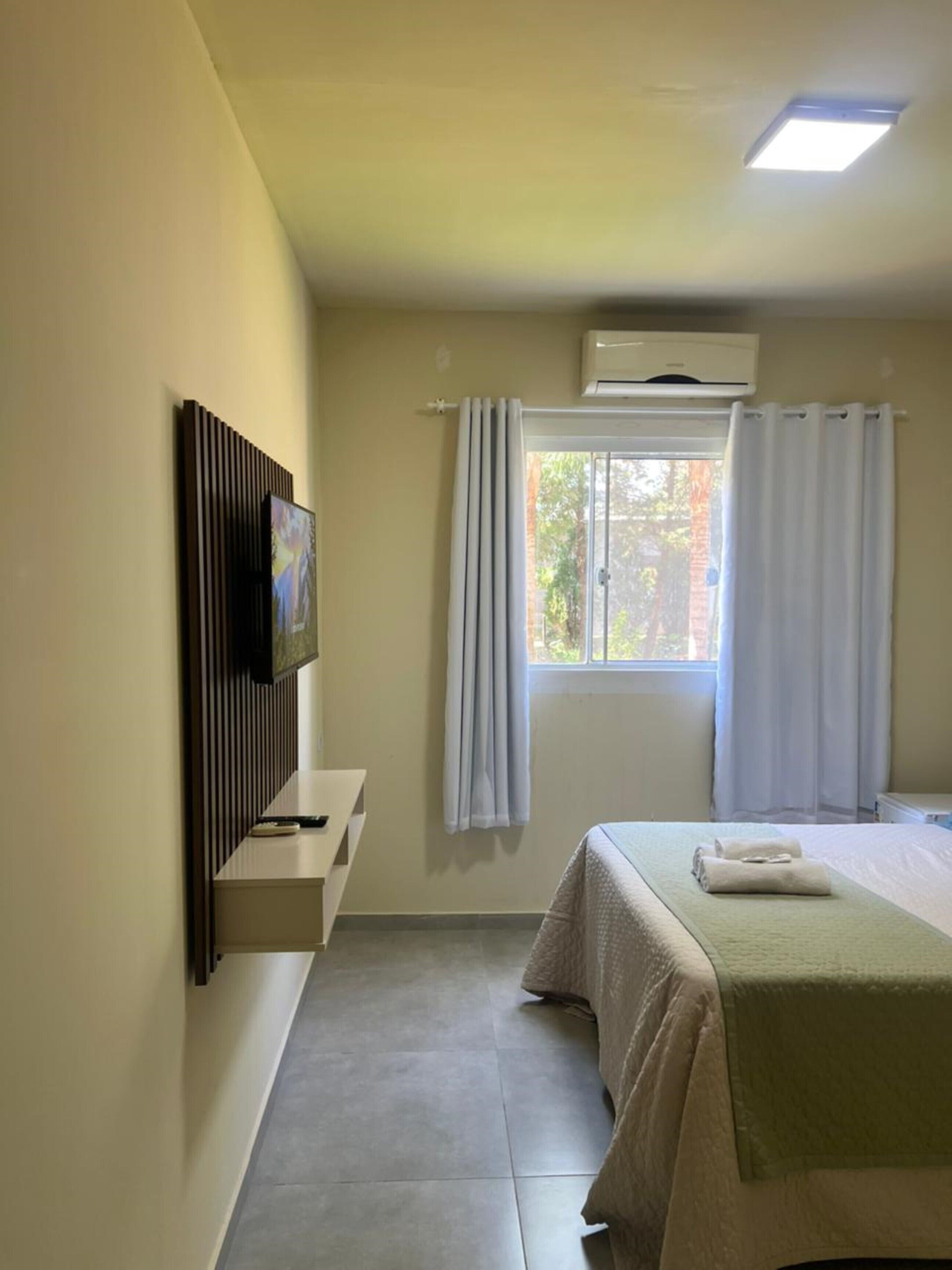 A hotel room with a bed, a flat-screen TV mounted on the wall, a window with curtains, and an air conditioning unit.
