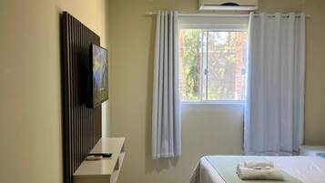 A hotel room with a bed, a flat-screen TV mounted on the wall, a window with curtains, and an air conditioning unit.