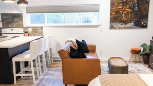 Una cocina moderna con una isla central, taburetes de bar blancos, un sofá marrón con cojines y un ventanal grande.