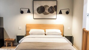 Un dormitorio con cama de madera, ropa de cama blanca, un cuadro abstracto colgado en la pared y dos lámparas de noche.
