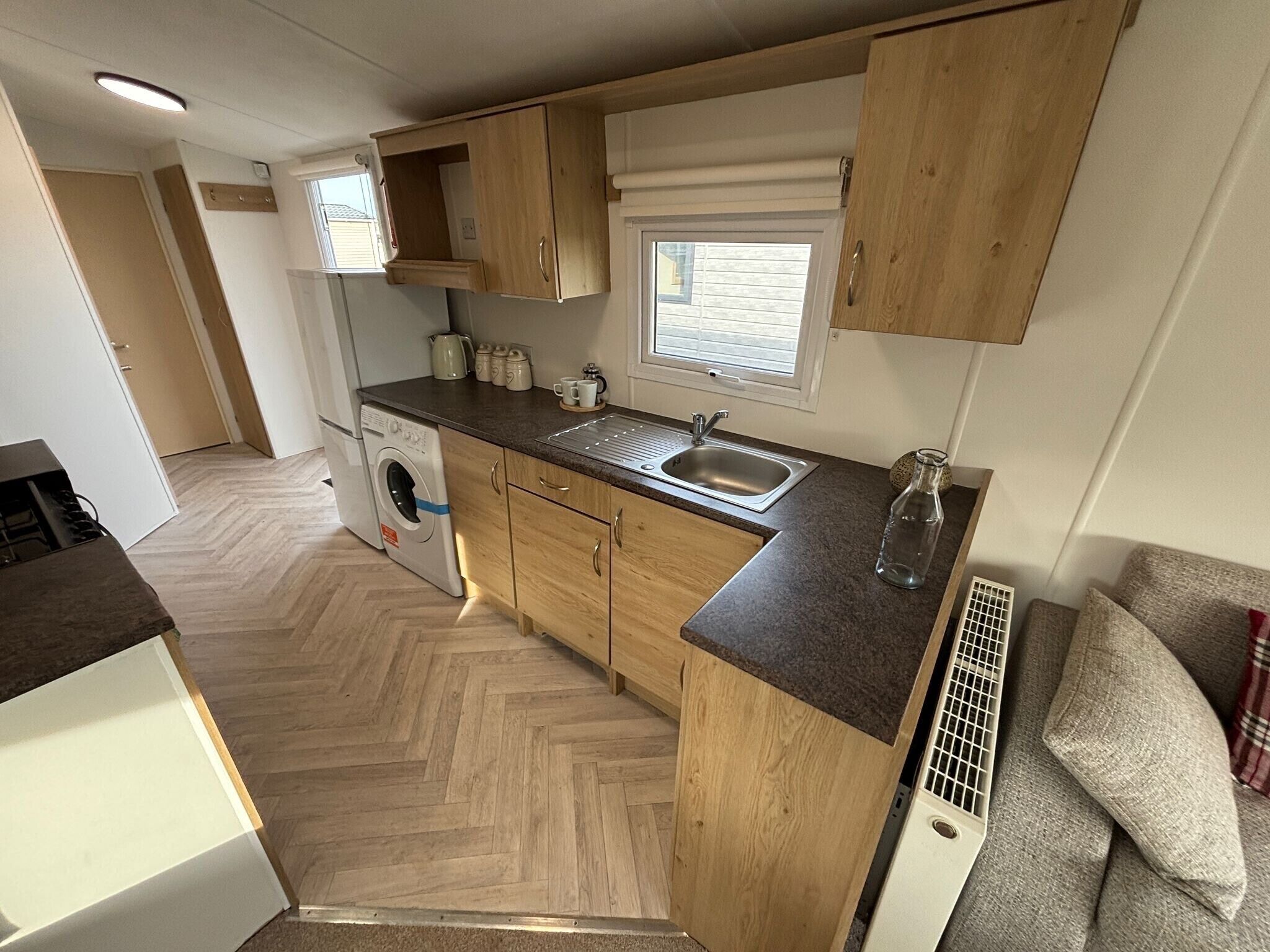 A compact kitchen with wooden cabinets, a built-in washing machine, a sink, and a window with blinds.