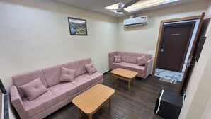 A living room with two sofas, a wooden coffee table, a ceiling fan, and an air conditioning unit. -  TRPOTELS THE ANANTA INN Near Old High Court Gwalior (Gwalior)