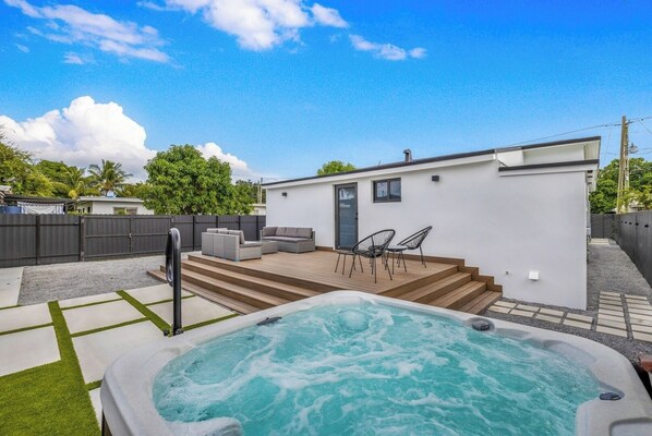 A modern outdoor hot tub area with a wooden deck, a small white building, and outdoor seating.