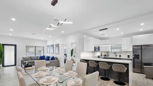 A modern kitchen with white cabinets, a stainless steel refrigerator, and a dining area with a glass table and chairs. - Design Dist Living:Pool,Basketball CourtandNespresso (Miami)