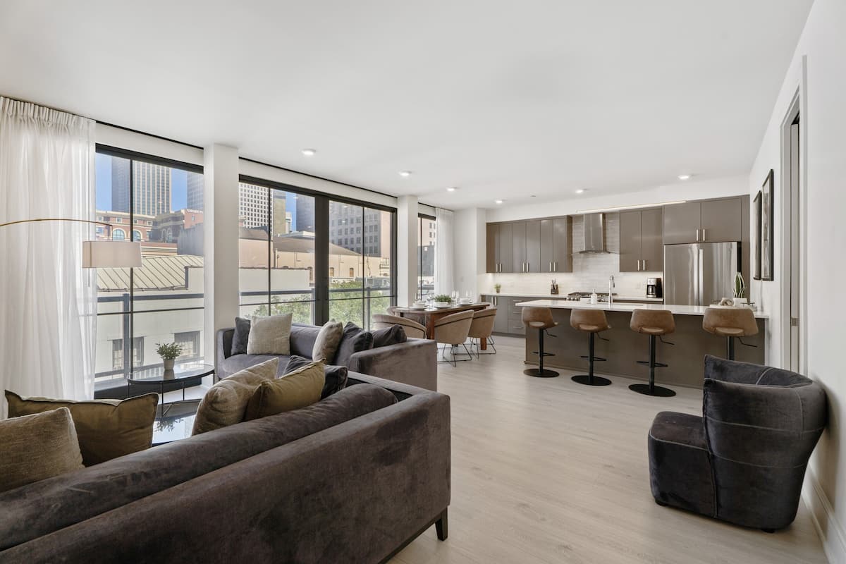 Una sala de estar moderna con un sofá y sillones, un comedor con mesa y sillas, y una cocina con taburetes de bar.