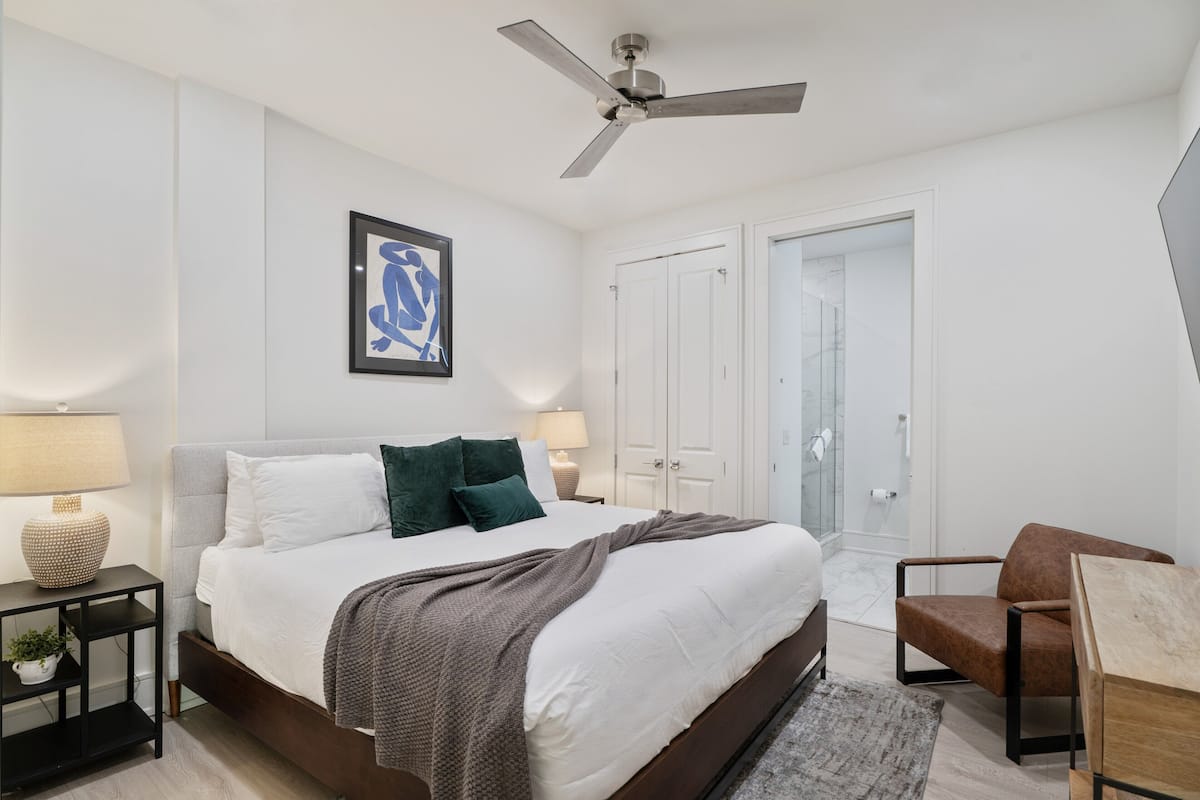 Un dormitorio moderno con cama, mesitas de noche, una silla, ventilador de techo y una obra de arte enmarcada en la pared.